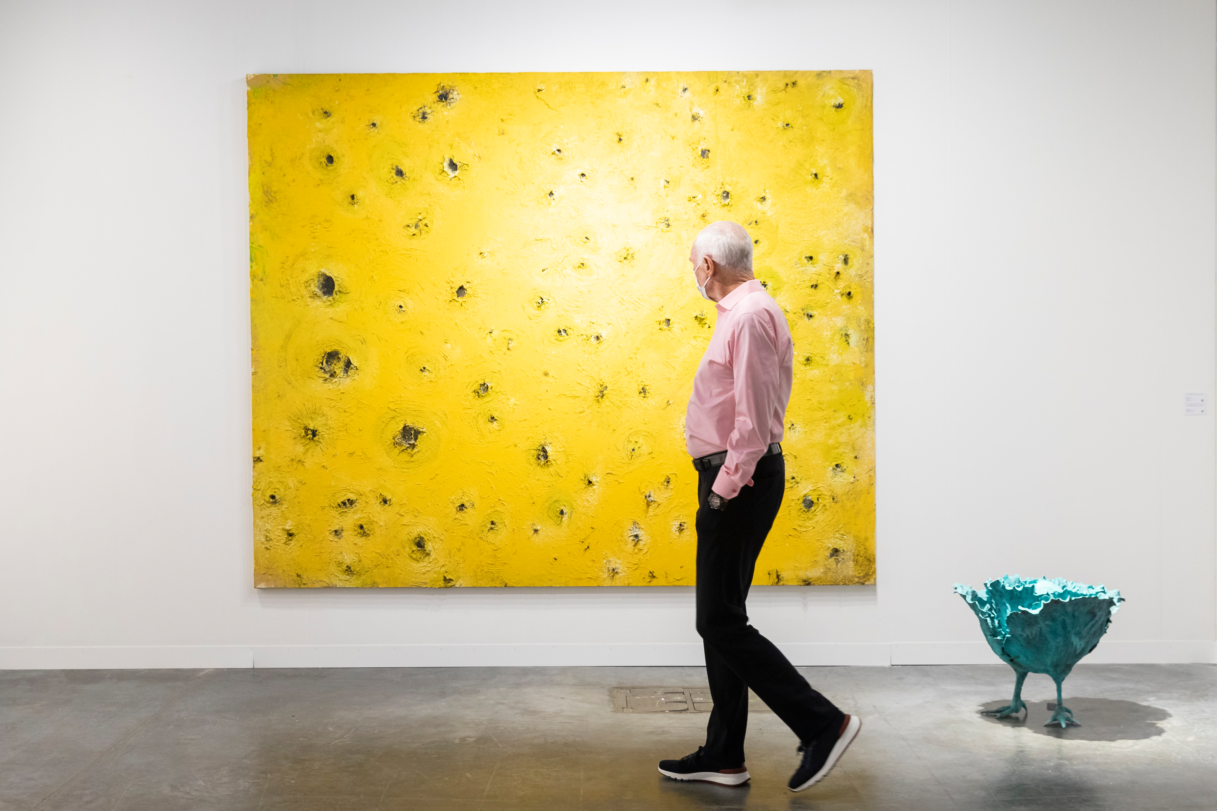 A man wearing a mask walks by a large yellow painting with black textured craters at Art Basel Miami Beach in 2021.